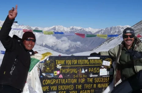 Thorong La Pass summit sign at 5,416 meters with colorful Buddhist prayer flags and snowy Annapurna mountain range in Nepal.