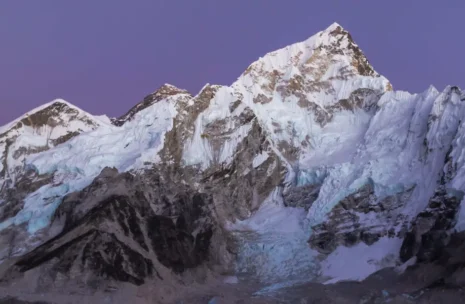 A long metal suspension bridge draped with colorful prayer flags spans a deep valley, leading toward steep pine-covered hills and massive snow-capped Himalayan peaks under a bright blue sky on the Everest Base Camp trek.
